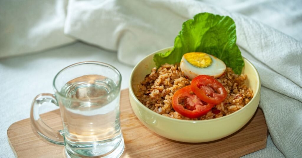 "A bowl of seasoned brown rice topped with a boiled egg, fresh tomato slices, and lettuce leaf beside a glass of water on a wooden board.
